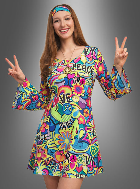 Eine Frau lächelt vor einem grauen Hintergrund mit Farbverlauf. Sie trägt das „Hippie Kleid Peace & Love“ – ein farbenfrohes, von den 1960er Jahren inspiriertes Kleid mit bunten Flower-Power-Mustern und ikonischen Peace-&-Love-Symbolen – und streckt beide Hände zum Peace-Zeichen aus.