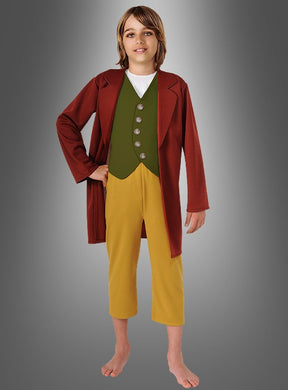 Ein Kind, das einen braunen Mantel, eine grüne Weste mit Knöpfen, eine gelbe Hose und keine Schuhe trägt, steht vor einem schlichten grauen Hintergrund mit Farbverlauf – ideal, um das Bilbo-Hobbit-Kinderkostüm oder jedes andere „Herr der Ringe“-Kostüm in Szene zu setzen.