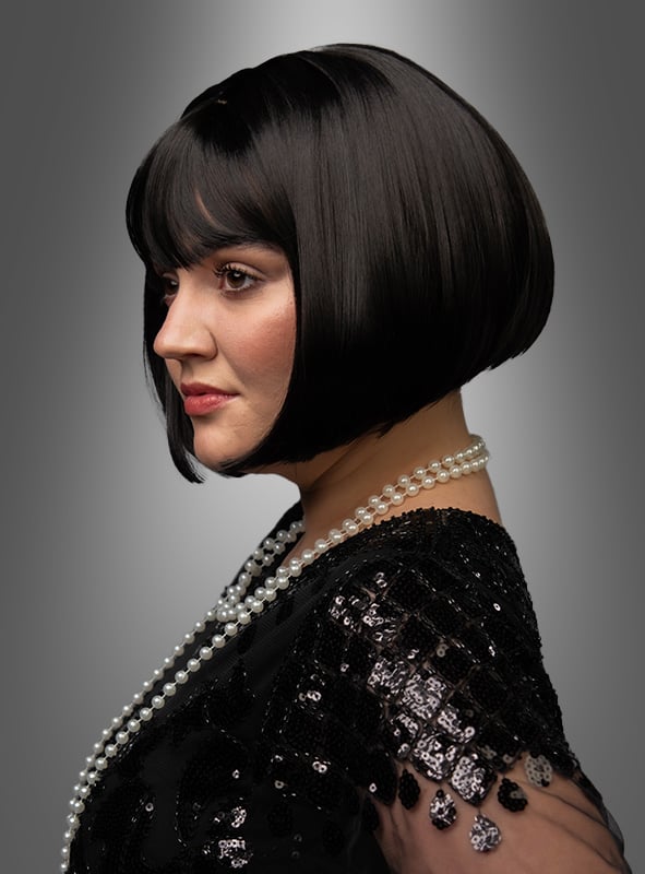 Eine Frau mit einem kurzen schwarzen Bob-Haarschnitt, die ein schwarzes Paillettenkleid und eine lange Perlenkette trägt, posiert im Profil vor einem grauen Hintergrund mit Farbverlauf. Ihr eleganter Look wird durch die stilvolle, hochwertige Bob-Perücke „Vera“ unterstrichen, die ihren raffinierten Stil perfekt ergänzt.