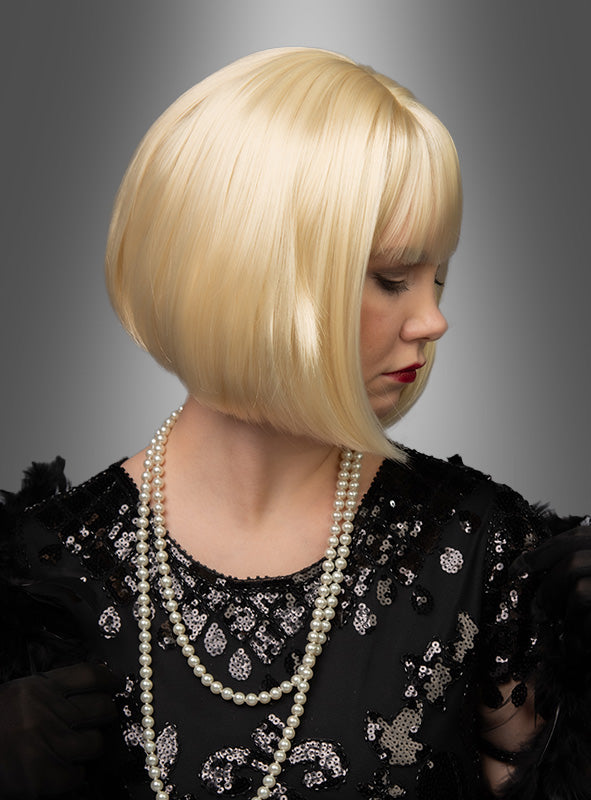 Eine Frau, die die hochwertige Pagenschnitt-Bob-Perücke „Vera“ in kurzem Platinblond trägt und mit einem schwarzen Pailletten-Oberteil, Perlenketten und schwarzen Handschuhen gestylt ist, blickt vor einem grauen Hintergrund mit Farbverlauf nach unten.