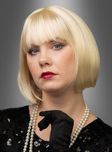Eine Frau präsentiert die hochwertige Pagenschnitt-Bob-Perücke „Vera“ mit kurzem blondem Haar im klassischen Bob-Stil. Sie trägt ein schwarzes Oberteil und schwarze Handschuhe, dazu eine Perlenkette und roten Lippenstift, und posiert vor einem grauen Hintergrund mit Farbverlauf.