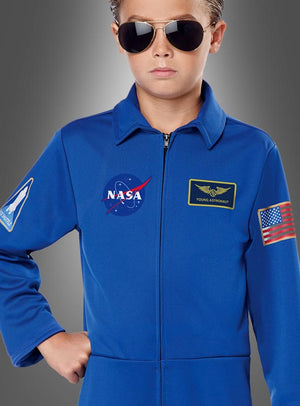 Ein Kind, das den blauen Astronautenanzug der NASA für Kinder trägt – mit Missionsabzeichen, Pilotenbrille und einem Aufnäher mit der amerikanischen Flagge –, steht vor einem grauen Hintergrund mit Farbverlauf.