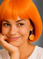 Eine lächelnde Person mit einem leuchtend orangefarbenen Bob-Haarschnitt trägt die „Orangen Ohrringe Scheibe“ – einen Ohrring in Form einer Orangenscheibe –, während sie ihr Kinn auf die Hand stützt; sie trägt ein weißes Hemd vor einem grauen Hintergrund.
