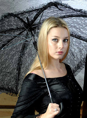 Eine Frau mit langen blonden Haaren, gekleidet in ein schwarzes Outfit, hält den Gothic-Schirm mit Spitze (weiß oder schwarz) in einem Innenraum in die Kamera und blickt dabei mit ausdruckslosem Gesicht in die Kamera – ein elegantes Ensemble, das sich perfekt für ein raffiniertes Halloween-Kostüm oder als stilvoller Sonnenschutz eignet.