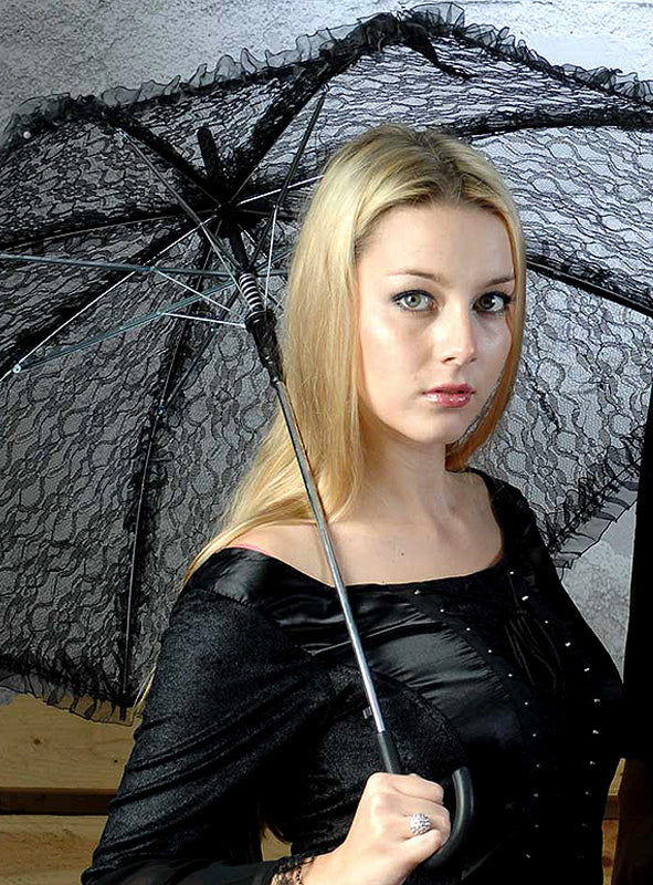 Eine Frau mit langen blonden Haaren, gekleidet in ein schwarzes Outfit, hält den Gothic-Schirm mit Spitze (weiß oder schwarz) in einem Innenraum in die Kamera und blickt dabei mit ausdruckslosem Gesicht in die Kamera – ein elegantes Ensemble, das sich perfekt für ein raffiniertes Halloween-Kostüm oder als stilvoller Sonnenschutz eignet.