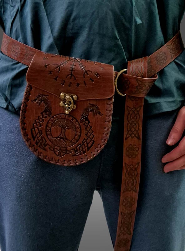 Eine Person, die eine braune Wikinger-Gürteltasche aus Leder mit Gürtel und aufwendigen Prägungen trägt, die über einer blauen Hose und einem grünlichen Oberteil befestigt ist – ein unverwechselbares Accessoire, das perfekt zu jedem Mittelalter-Kostüm passt.