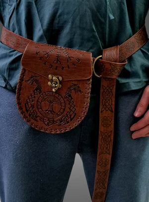 Eine Person, die eine braune Wikinger-Gürteltasche aus Leder mit Gürtel und aufwendigen Prägungen trägt, die über einer blauen Hose und einem grünlichen Oberteil befestigt ist – ein unverwechselbares Accessoire, das perfekt zu jedem Mittelalter-Kostüm passt.