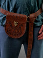 Eine Person, die eine braune Wikinger-Gürteltasche aus Leder mit Gürtel und aufwendigen Prägungen trägt, die über einer blauen Hose und einem grünlichen Oberteil befestigt ist – ein unverwechselbares Accessoire, das perfekt zu jedem Mittelalter-Kostüm passt.