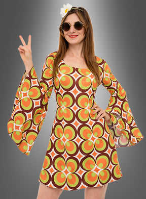 Eine Frau, die das waschbare 70er-Retro-Kleid „Nicky“ trägt, dazu eine runde Sonnenbrille und ein Blumenstirnband, zeigt vor einem grauen Hintergrund mit Farbverlauf das Peace-Zeichen.