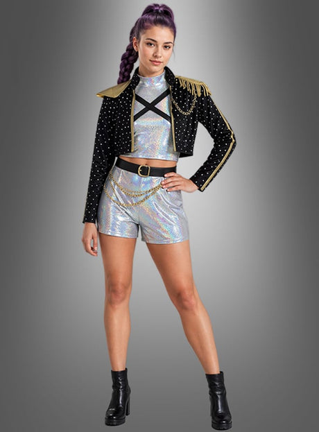 Eine Person mit lila Haaren, die zu einem geflochtenen Pferdeschwanz zusammengebunden sind, trägt das K-Pop-Star-Kostüm „Romy Sängerin“ – ein glitzerndes silbernes Outfit –, kombiniert mit einer schwarzen Nietenjacke mit goldenen Verzierungen und schwarzen Stiefeletten, und steht vor einem grauen Hintergrund.