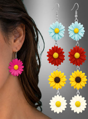 Nahaufnahme einer Frau, die einen rosa „Blumen Ohrringe Flower Power“-Ohrring trägt, der den Boho-Look verkörpert, zusammen mit vier weiteren bunten „Blumen Ohrringe Flower Power“-Ohrringen – in Blau, Rot, Gelb und Weiß –, die rechts vor einem grau verlaufenden Hintergrund zu sehen sind.