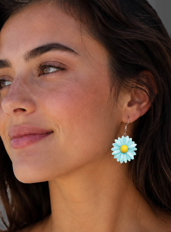 Eine Frau mit langen, dunklen Haaren, die sanft lächelt, während sie zur Seite blickt, trägt die „Blumen Ohrringe Flower Power“ – hellblaue und gelbe Ohrringe in Gänseblümchenform, die den Boho-Look verkörpern.