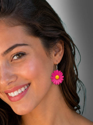 Eine Frau mit langen braunen Haaren lächelt und trägt die farbenfrohen „Blumen Ohrringe Flower Power“ – in Form von Gänseblümchen in leuchtendem Rosa und Gelb –, die den verspielten Geist von Flower Power einfangen und für einen fröhlichen Boho-Look sorgen.