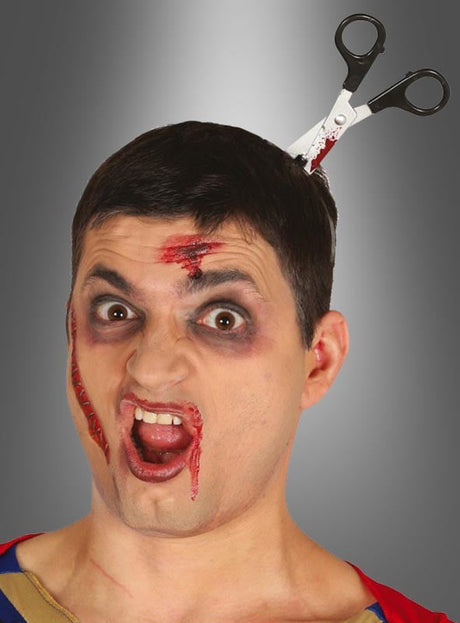 Ein Mann mit theatralischem Make-up, das Wunden vortäuscht, und einem Stirnband mit der Aufschrift „Horror Haarreif Werkzeuge im Kopf“, auf dem eine künstliche Schere zu sehen ist, die scheinbar seinen Kopf durchbohrt, posiert mit schockiertem Gesichtsausdruck.