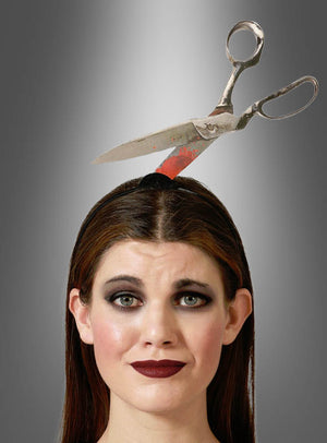 Eine Frau mit dunklem Make-up trägt das Halloween-Accessoire „Horror Haarreif Werkzeuge im Kopf“, ein Stirnband, das so gestaltet ist, dass es aussieht, als stecke eine große Schere durch ihren Kopf; im Hintergrund ist ein grauer Farbverlauf zu sehen.