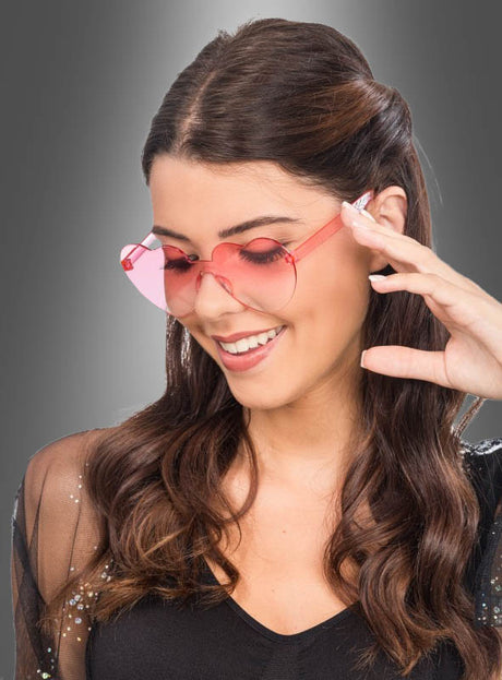 Eine Frau mit langen braunen Haaren, die ein schwarzes Oberteil trägt und die rosa „Herzbrille“-Karnevals-Sonnenbrille für Kostümpartys aufhat, lächelt, während sie ihre Brille vor einem grauen Farbverlaufshintergrund in die Hand nimmt – ein ideales Accessoire für den Karnevalsspaß.