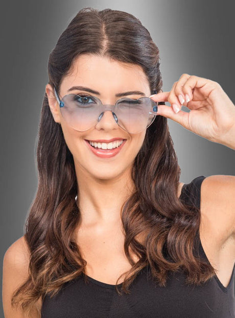 Eine Frau mit langen braunen Haaren, die ein schwarzes Oberteil trägt, lächelt, während sie ihre stilvolle Sonnenbrille „Herzbrille Karneval zum Kostüm“ mit einer Hand vor einem grauen Farbverlaufshintergrund hochschiebt.