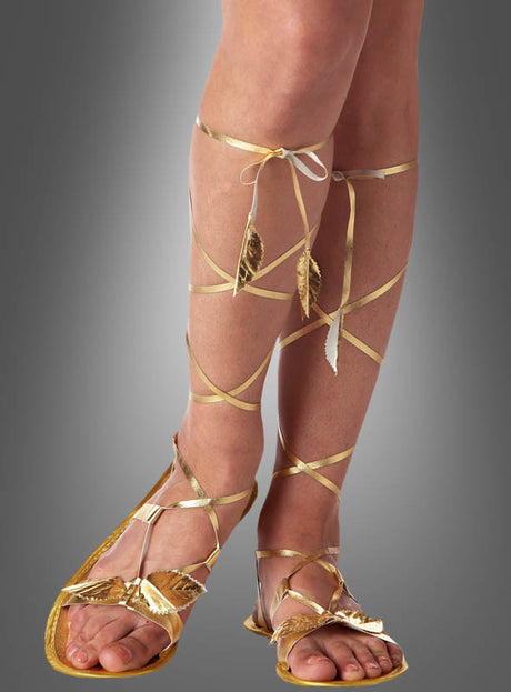 Jemand, der „Göttin“-Sandalen trägt – goldene Sandalen, die von Kleopatra-Schuhen inspiriert sind, mit Blattmotiven und langen, sich kreuzenden Riemen, die sich um die Unterschenkel wickeln und dort zusammengebunden werden – perfekt für ein „Göttin“-Kostüm, posiert vor einem schlichten Hintergrund mit Farbverlauf.