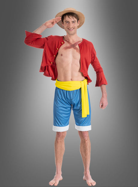 Ein Mann mit Strohhut, offenem rotem Hemd, gelber Schärpe und blauen Shorts steht barfuß vor einem grauen Hintergrund und salutiert lächelnd – perfekt für alle, die nach dem Ruffy-Kostüm für Erwachsene aus „One Piece“ suchen.