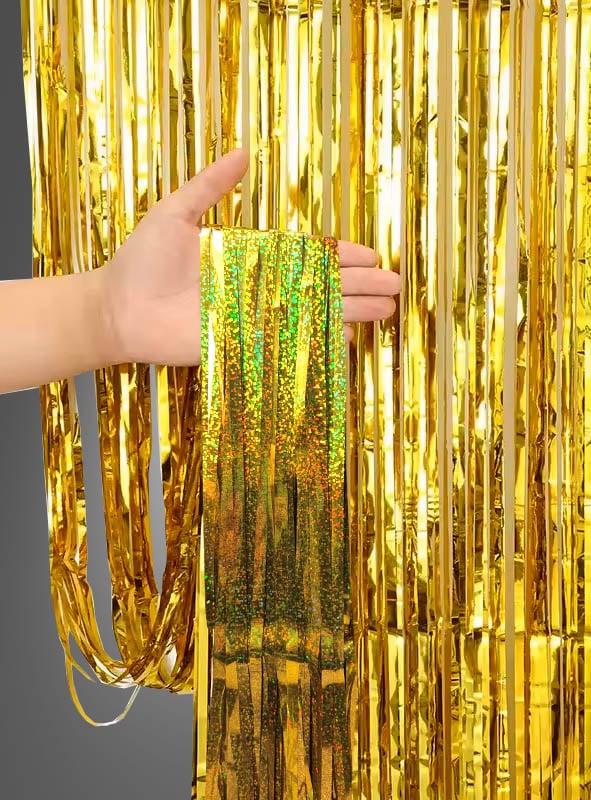 Eine Hand hält einen Abschnitt des „Glitter Lametta Vorhangs“ (100 x 250 cm) hoch – einen glänzenden, goldfarbenen Metallic-Fransenvorhang, der sich perfekt als Glitzervorhang für die Partydekoration eignet – vor dem Hintergrund ähnlich hängender goldener Streifen.