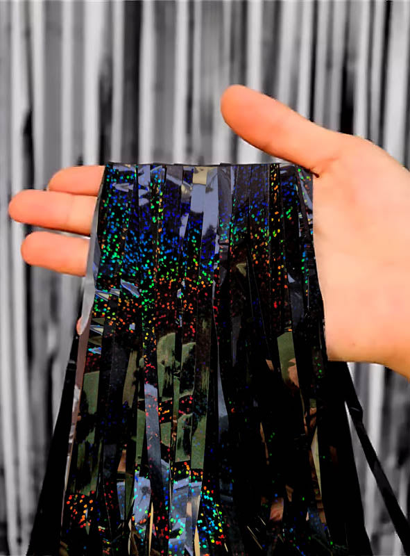 Eine Hand hält schwarze und holografische Lamettafäden vor einem schwarz-weiß gestreiften Lametta-Hintergrund – ideal, um einen auffälligen „Glitzervorhang“-Partydeko zu gestalten oder als stilvollen „Glitter-Lametta-Vorhang“ (100 x 250 cm) für jede Feier.
