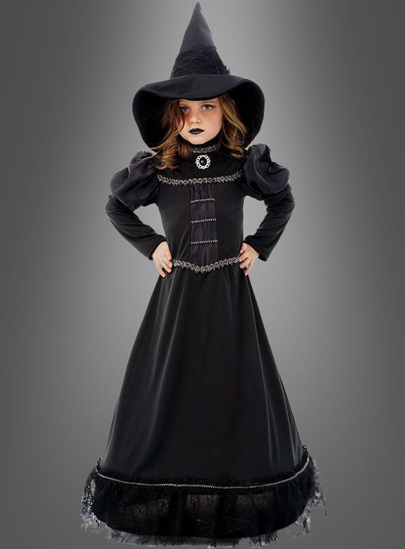 Ein junges Mädchen trägt das „Deluxe schwarze Hexe Moyra“-Kinderkostüm, bestehend aus einem langen schwarzen Hexenoutfit und einem spitzen Hut, und steht mit den Händen in die Hüften gestemmt vor einem grauen Hintergrund.