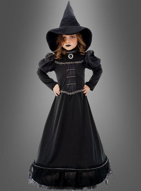 Ein junges Mädchen trägt das „Deluxe schwarze Hexe Moyra“-Kinderkostüm, bestehend aus einem langen schwarzen Hexenoutfit und einem spitzen Hut, und steht mit den Händen in die Hüften gestemmt vor einem grauen Hintergrund.