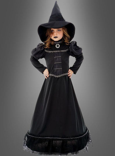 Ein junges Mädchen trägt das „Deluxe schwarze Hexe Moyra“-Kinderkostüm, bestehend aus einem langen schwarzen Hexenoutfit und einem spitzen Hut, und steht mit den Händen in die Hüften gestemmt vor einem grauen Hintergrund.