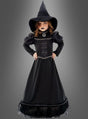 Ein junges Mädchen trägt das „Deluxe schwarze Hexe Moyra“-Kinderkostüm, bestehend aus einem langen schwarzen Hexenoutfit und einem spitzen Hut, und steht mit den Händen in die Hüften gestemmt vor einem grauen Hintergrund.