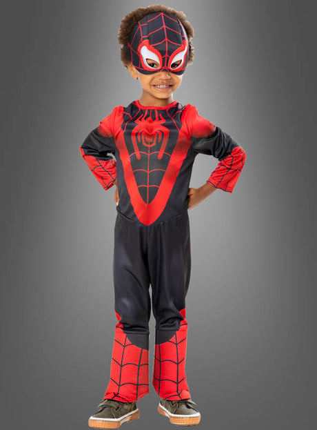 Ein Kind, das das schwarz-rote Spiderman-Kinderkostüm trägt, steht lächelnd mit den Händen in den Hüften vor einem schlichten grauen Hintergrund.