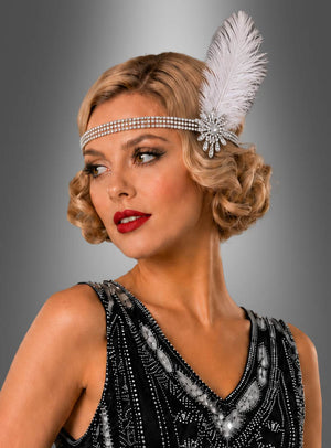 Eine Frau mit kurzen blonden Locken trägt den „20er Strass Kopfschmuck mit Federn“ – ein mit Perlen verziertes Flapper-Stirnband mit einer weißen Feder – sowie roten Lippenstift und ein schwarz-silbernes Kleid im Stil der 1920er Jahre; sie blickt vor einem grauen Hintergrund nach links.