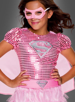 Ein Kind, das das „Rosa Supergirl“-Kleid trägt – ein glänzendes rosa Superheldenkostüm mit einem S-Emblem, dazu eine passende rosa Maske, ein Umhang und ein Tutu –, steht mit den Händen in die Hüften gestemmt vor einem grauen Hintergrund.