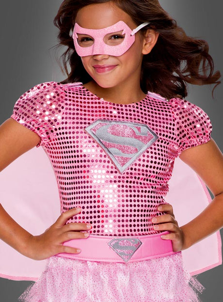Ein Kind, das das „Rosa Supergirl“-Kleid trägt – ein glänzendes rosa Superheldenkostüm mit einem S-Emblem, dazu eine passende rosa Maske, ein Umhang und ein Tutu –, steht mit den Händen in die Hüften gestemmt vor einem grauen Hintergrund.