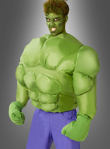 Eine Person trägt das aufblasbare Hulk-Kostüm für Herren mit lila Hose und posiert mit geballten Fäusten wie ein Comic-Superheld vor einem grauen Hintergrund mit Farbverlauf.