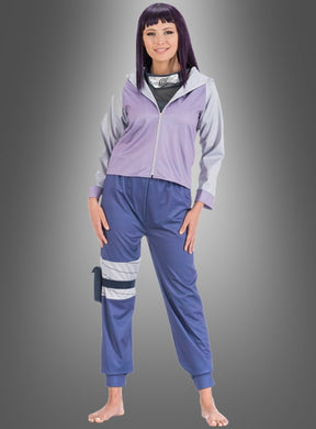 Eine Person steht barfuß und lächelt. Sie trägt ein lila Hinata-Kostüm für Erwachsene aus Naruto mit einer Jacke, einem Ninja-Stirnband und Schienbeinschützern vor einem schlichten grauen Hintergrund.