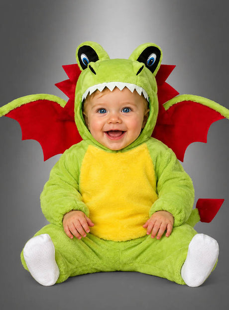 Ein Baby, das das „Kuscheliges Drachen“-Babykostüm in Grün und Gelb mit roten Flügeln trägt, sitzt vor einem grauen Hintergrund und lächelt.