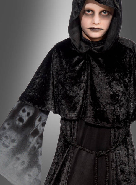 Eine Person, die das Halloween-Kostüm „Vergessene Seelen“ trägt – ein schwarzes Samtgewand mit Geistergesichtern an den Ärmeln –, dazu blasses Make-up sowie dunkles Augen- und Lippen-Make-up vor grauem Hintergrund: das perfekte Halloween-Kostüm, um vergessene Seelen zu verkörpern.