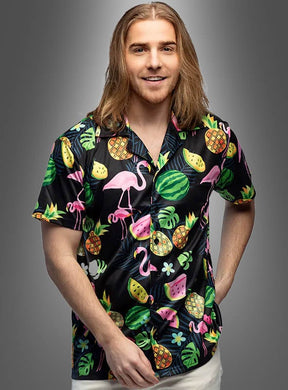 Ein Mann mit langen Haaren trägt das Hawaiihemd „Flamingo Style Herren“ – ein schwarzes Hemd, das mit bunten Flamingos, Wassermelonen, Ananas und tropischen Blättern verziert ist – und steht vor einem grauen Hintergrund.