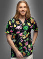Ein Mann mit langen Haaren trägt das Hawaiihemd „Flamingo Style Herren“ – ein schwarzes Hemd, das mit bunten Flamingos, Wassermelonen, Ananas und tropischen Blättern verziert ist – und steht vor einem grauen Hintergrund.