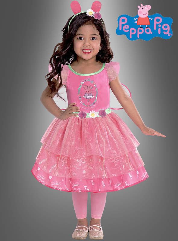 Ein kleines Mädchen, das das „Peppa Pig“-Feenkleid für Mädchen trägt – bestehend aus einem rosa Kleid mit Peppa-Wutz-Motiv, passenden rosa Leggings und einem Stirnband mit Ohren –, steht lächelnd vor einem grauen Hintergrund, auf dem das Peppa-Pig-Logo zu sehen ist.