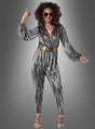 Eine Frau im Disco-Kostüm-Jumpsuit „Super Nova“, kombiniert mit einem metallisch gestreiften Muster, einem goldenen Gürtel und einer Sonnenbrille, steht lächelnd vor einem grauen Hintergrund – und verkörpert damit perfekt das ultimative „Dancing Queen“-Outfit.