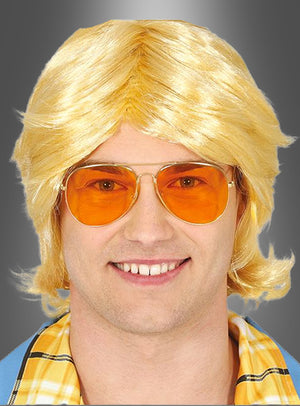 Ein Mann mit einer blonden Perücke, einer Retro-Brille aus den 70er Jahren mit orange getönten Pilotenbrillengläsern und einem gelben Karohemd lächelt vor einem grauen Hintergrund mit Farbverlauf in die Kamera.
