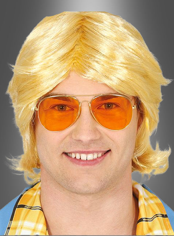 Ein Mann mit einer blonden Perücke, einer Retro-Brille aus den 70er Jahren mit orange getönten Pilotenbrillengläsern und einem gelben Karohemd lächelt vor einem grauen Hintergrund mit Farbverlauf in die Kamera.