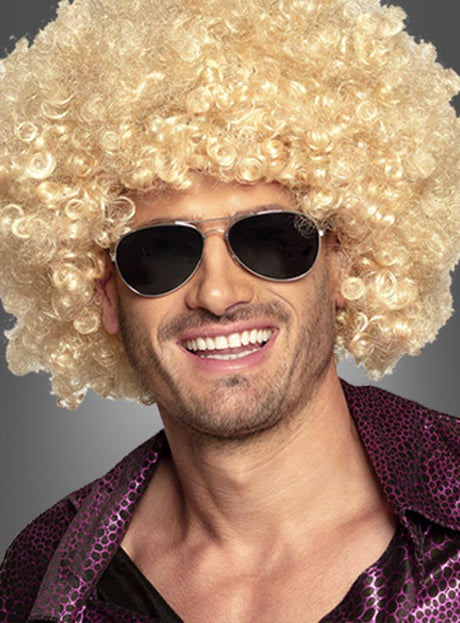Ein Mann mit einer großen, lockigen blonden Perücke, einer Sonnenbrille im Retro-Stil der 70er Jahre und einem lila gemusterten Hemd lächelt vor einem grauen Hintergrund in die Kamera – perfekt für jedes Kostümzubehör oder jede Party im Stil der 70er Jahre.