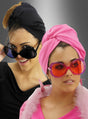 Zwei Frauen mit stilvollen Kopftüchern und großen Retro-Brillen mit großen Gläsern – die eine in Schwarz mit passender schwarzer Brille, die andere in Rosa mit einer verspielten rosa Retro-Brille mit großen Gläsern und einem Federkragen – stehen selbstbewusst vor einem grauen Hintergrund mit Farbverlauf.