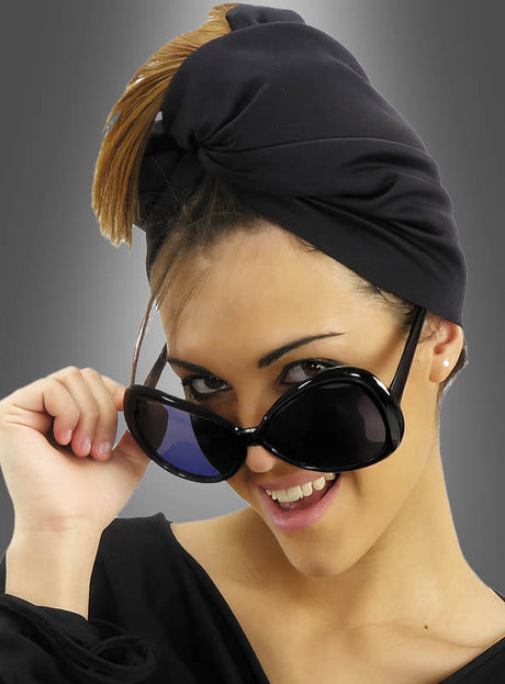 Eine Frau mit schwarzem Kopftuch und einer großen Retro-Sonnenbrille mit großen Gläsern lächelt, während sie die Sonnenbrille leicht anhebt; im Hintergrund ist ein grauer Farbverlauf zu sehen.