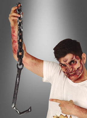 Ein Mann mit theatralischem Gesichts-Make-up und künstlichen Wunden, gekleidet in ein Henker-Kostüm, hält die große, schwarze Requisite „Ketten mit blutigen Fleischerhaken“ hoch und zeigt darauf; der grau abgestufte Hintergrund macht diese Szene zur perfekten Inspiration für Ihre Halloween-Dekoration.