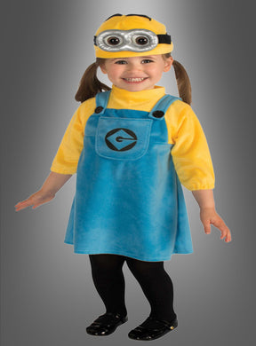 Ein kleines Kind, das das Original-Minion-Mädchen-Kostüm trägt – bestehend aus einem blauen Strickkleid, einem gelben Shirt, schwarzen Strumpfhosen, schwarzen Schuhen und einer Minion-Augenmaske –, steht lächelnd vor einem grauen Hintergrund.