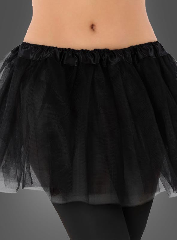 Vor einem grauen Hintergrund ist eine Person zu sehen, die den „Tütü Kinder Tüllrock“ trägt, einen kurzen schwarzen Tutu-Rock, kombiniert mit schwarzen Leggings und einem bauchfreien Oberteil.