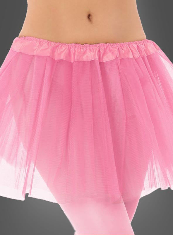Eine Person, die den rosa Tutu-Rock „Tütü Kinder Tüllrock“ und eine rosa Strumpfhose trägt, vor einem grauen Hintergrund. Das Bild ist so beschnitten, dass nur der untere Teil des Rumpfes und die Oberschenkel zu sehen sind.